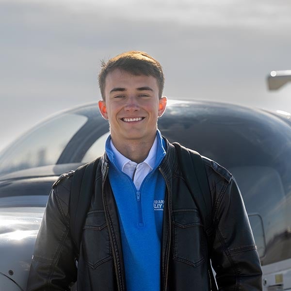 Smiling young man wearing a blue sweater and black jacket stands in front of a small airplane on an airport tarmac.”