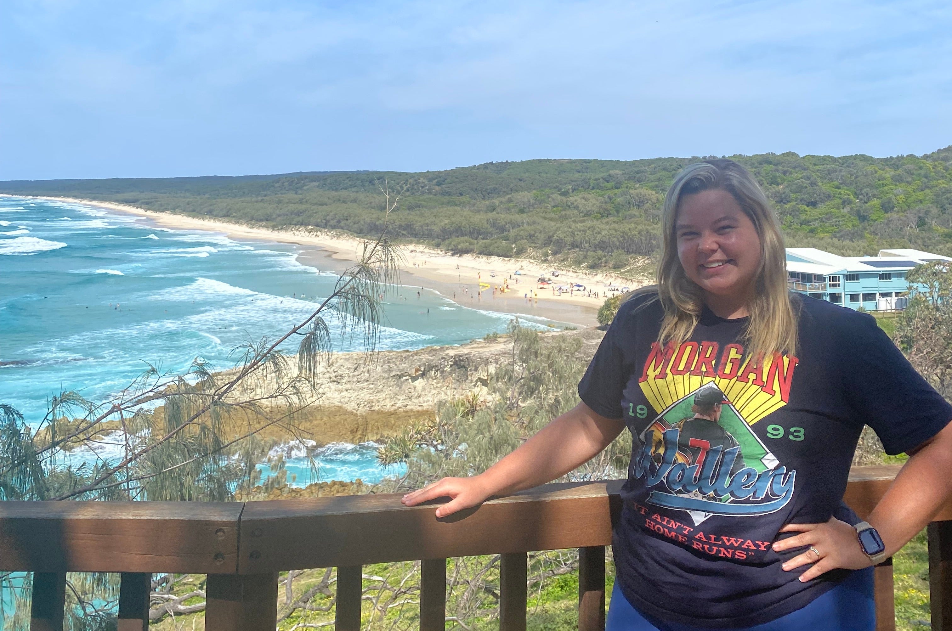 A white female stands at an overlook railing above a long sandy beach, with turquoise ocean waves, coastal cliffs, and dunes stretching along the shoreline. 