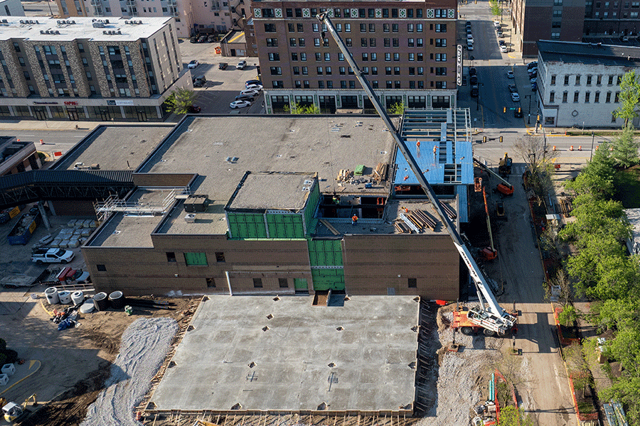 Aerial daytime view of a multi-story building under construction in an urban area, with a crane extending over the structure. Construction materials, scaffolding, and workers are visible on the site, surrounded by nearby streets and completed buildings.