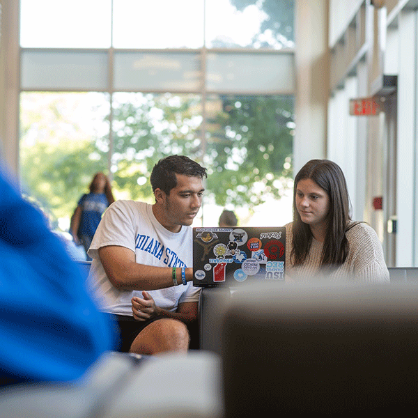 Two students sit together in a bright campus atrium, collaborating on a laptop covered with stickers, with large windows, seating, and other students visible in the background. 