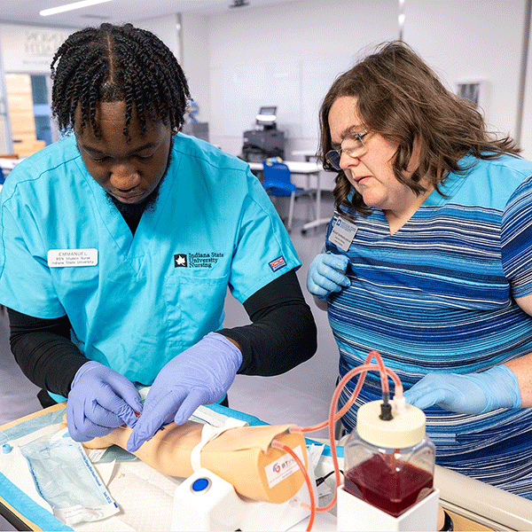 Two people in a clinical training classroom practice a medical procedure using a simulated arm, wearing gloves while working with tubing, medical supplies, and a blood collection container on a table.
