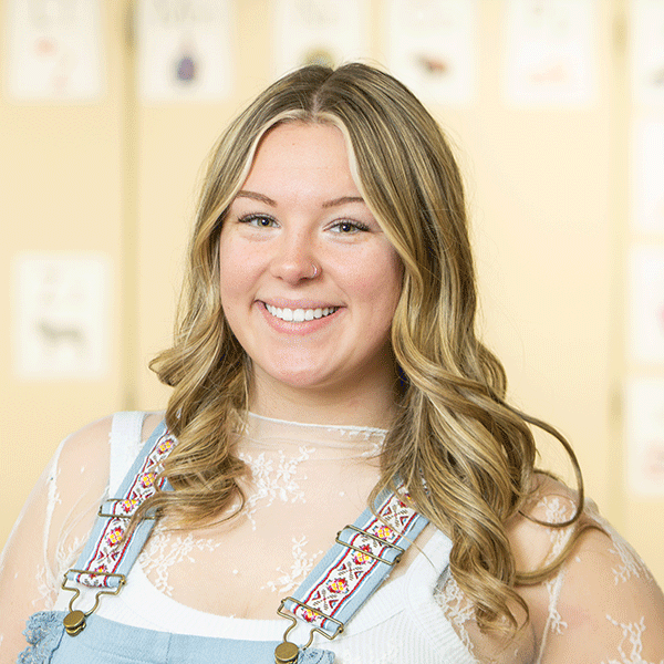 Smiling young woman with long, wavy blonde hair and wearing a sheer lace top, a white tank top, and light blue overalls with patterned straps, stands in front of a softly lit wall with student artwork taped to the wall behind her.