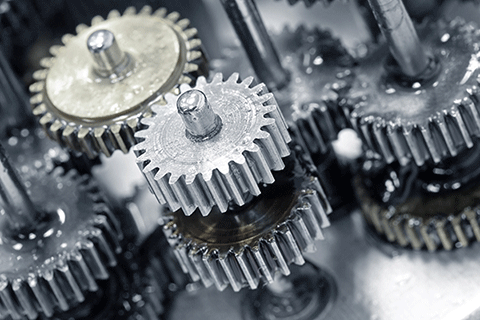 Close-up view of interlocking metal gears coated with lubricant, showing multiple toothed wheels mounted on shafts inside a mechanical assembly. The image emphasizes precision components and motion within machinery.