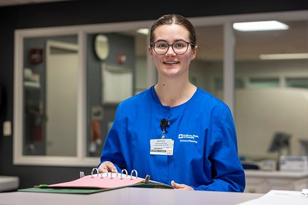 Young woman with glasses and dark hair pulled back and wearing blue medical scrubs with an ID badge, standing at a counter and holding an open binder, in a clinical workspace with interior windows and office equipment visible in the background.
