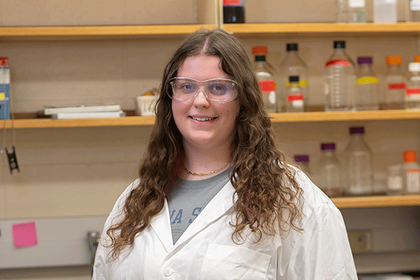 Person wearing a white lab coat and clear safety goggles, smiling while standing in a laboratory with shelves of labeled bottles and equipment in the background.