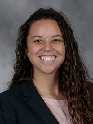 Head-and-shoulders portrait of a smiling woman with long, dark curly hair, wearing a black blazer over a light top, against a gray studio background.