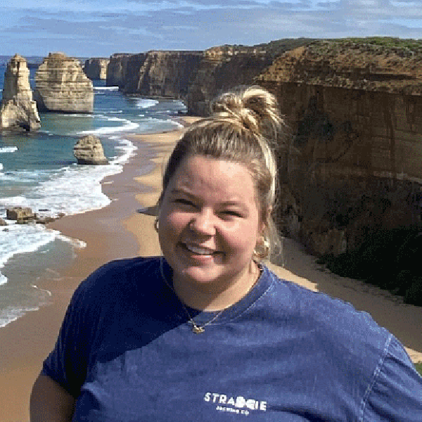 A woman stands smiling near a coastal cliff overlooking the ocean, with sandy shoreline, rolling waves, and tall limestone rock formations extending along the coastline beneath a bright, partly cloudy sky.