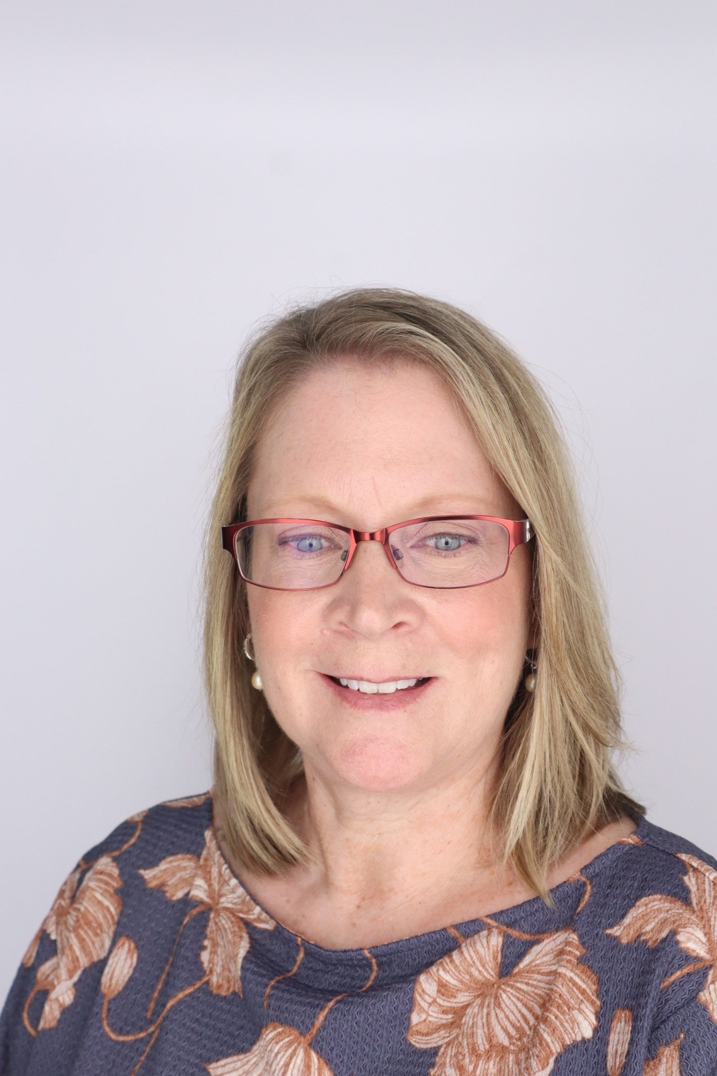 Head-and-shoulders portrait of a smiling woman with shoulder-length blonde hair, wearing red-framed glasses and a patterned top, against a plain light background.