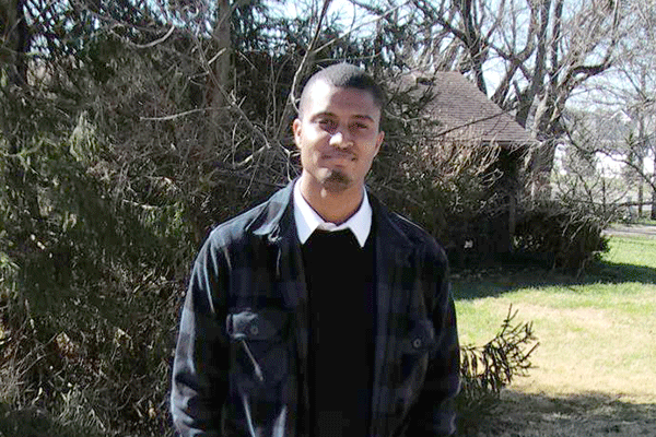 A person stands outdoors in a yard on a sunny day, wearing a dark plaid jacket over a black sweater and white collared shirt. Behind them are leafless trees, a wooden house with a sloped roof, and patches of grass lit by natural light.
