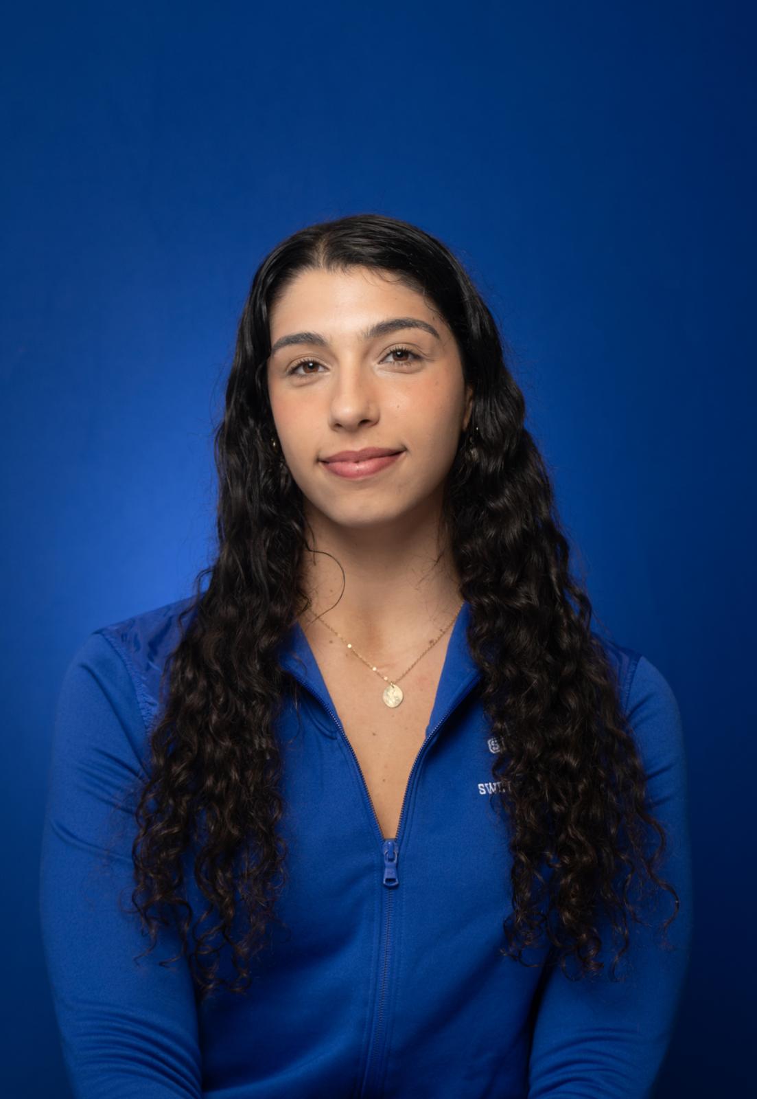Head-and-shoulders portrait of a woman with long, dark curly hair wearing a blue zip-up top and a necklace, against a solid blue background.