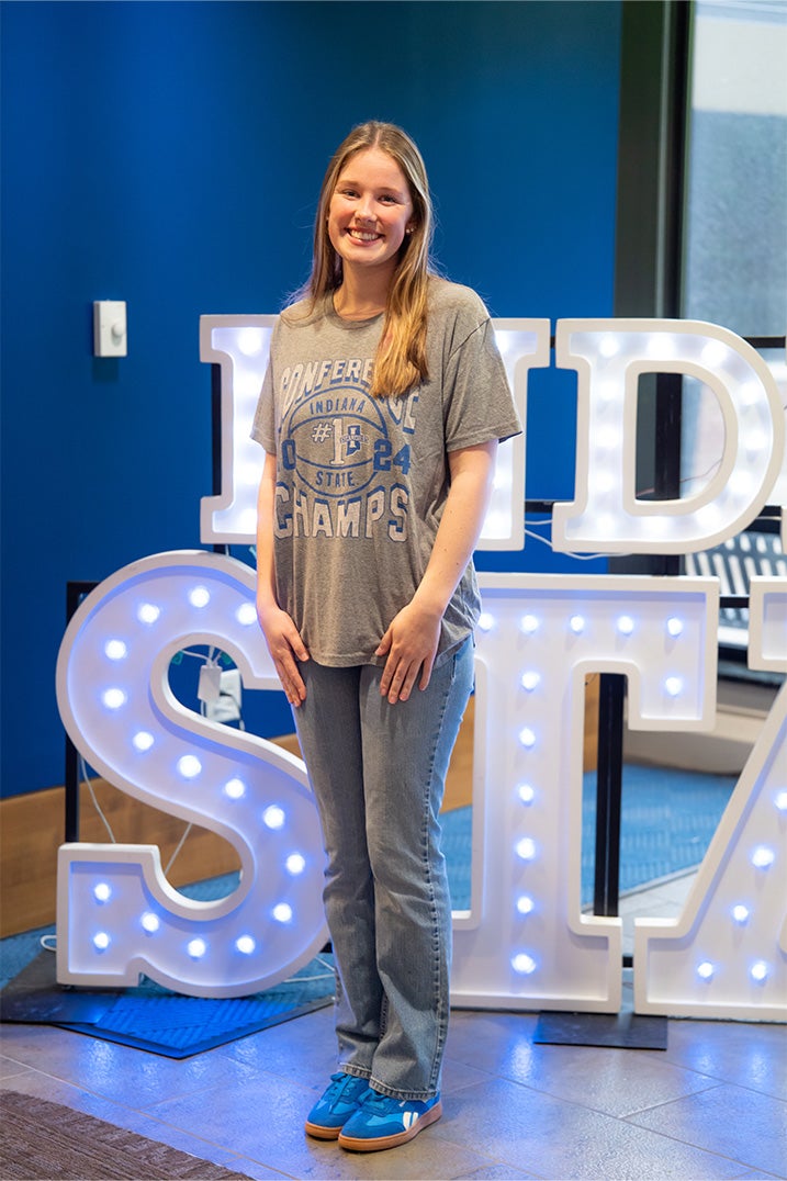 A smiling person with long blonde hair stands indoors in front of illuminated “Indiana State” display letters. They are wearing a gray Indiana State conference champions T-shirt, light blue jeans, and bright blue sneakers. Blue walls and large windows are visible in the background.