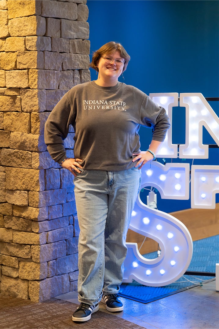 A smiling person with short light brown hair and glasses stands indoors beside a stone wall and illuminated “Indiana State” display letters. They are wearing a gray Indiana State University long-sleeve shirt, light blue jeans, and black sneakers with white stripes, with their hands on their hips. Blue walls and lit signage are visible in the background.
