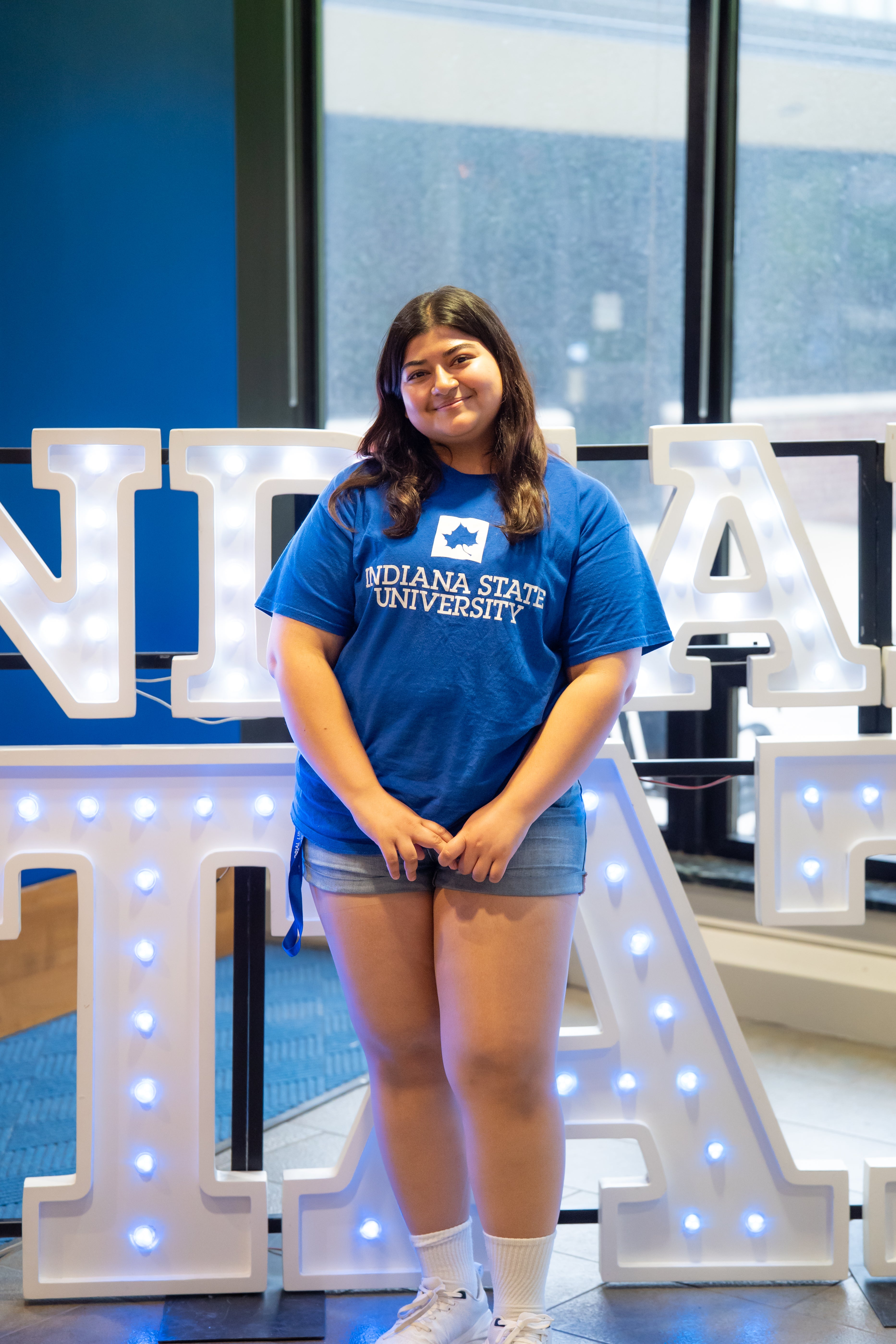 A smiling person with long dark hair stands indoors in front of illuminated “Indiana State” display letters and large windows. They are wearing a bright blue Indiana State University T-shirt, denim shorts, white crew socks, and white sneakers, with their hands clasped in front of them. Blue walls and lit signage are visible in the background.