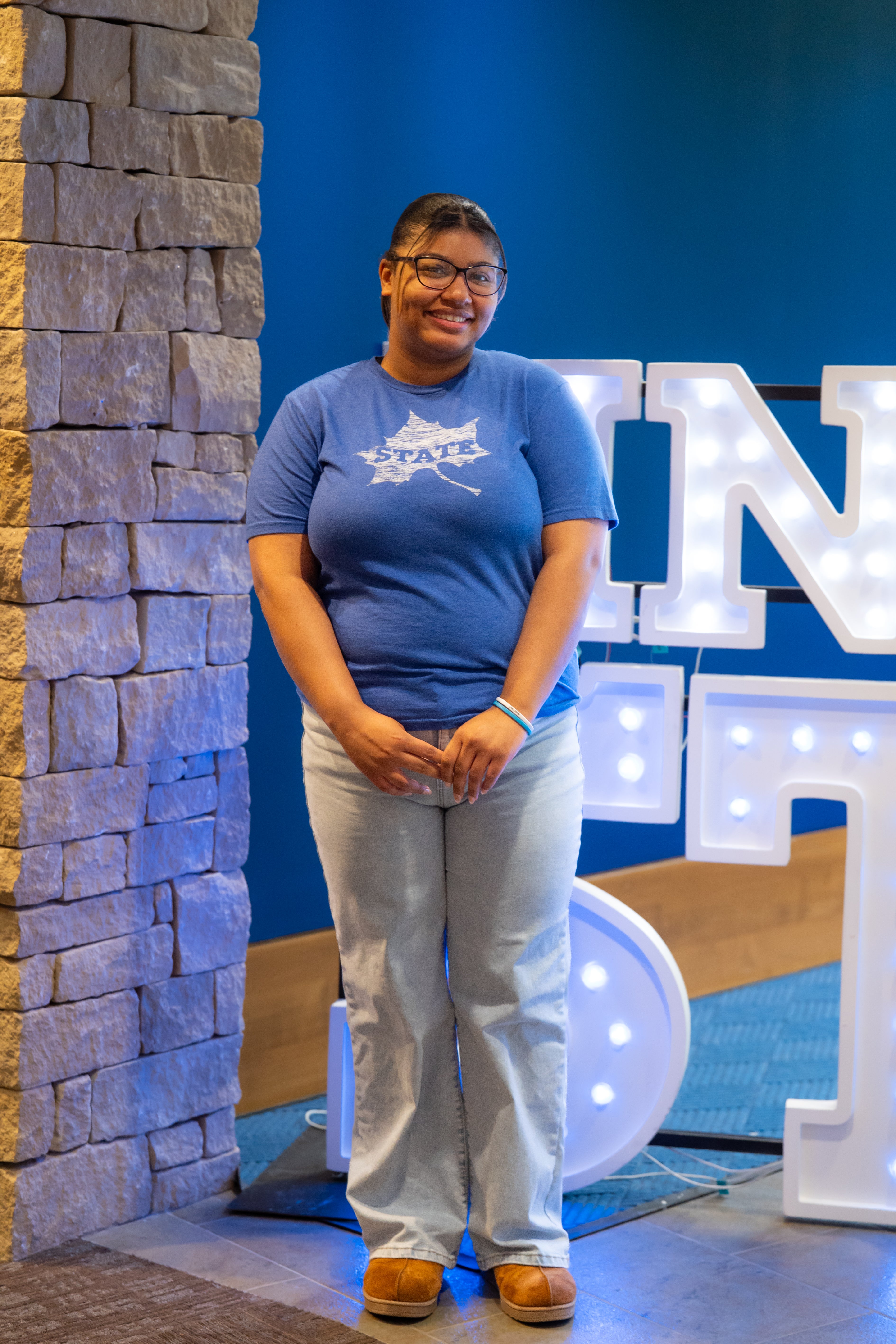 A smiling person with dark hair pulled back and glasses stands indoors beside a stone wall and illuminated “Indiana State” display letters. They are wearing a bright blue Indiana State T-shirt, light blue jeans, and tan shoes, with their hands clasped in front of them. Blue walls and lit signage are visible in the background.