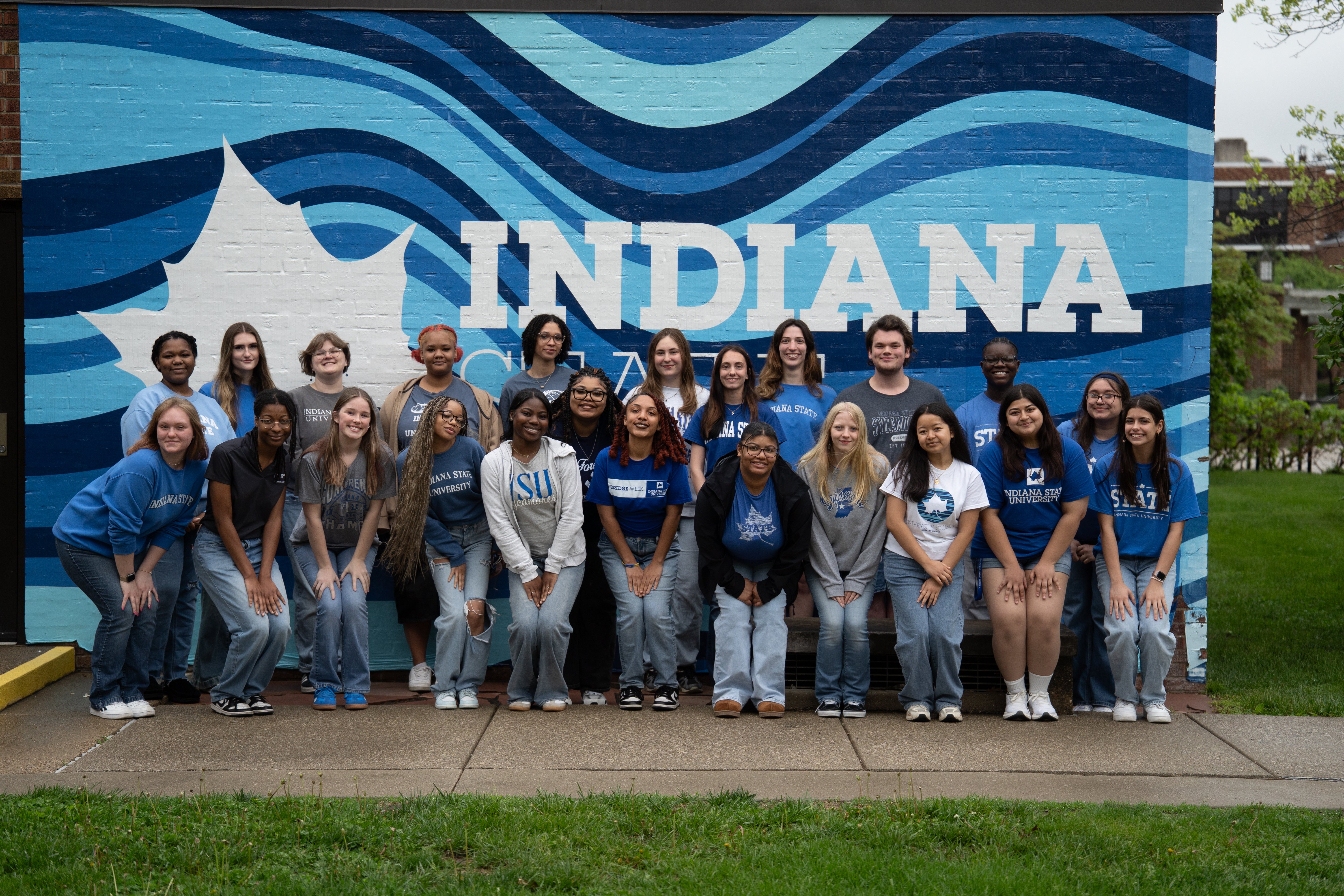 A group of students stands and kneels together in front of a large blue-and-white Indiana State mural outdoors. The mural features a white sycamore leaf and large “INDIANA” lettering over layered wave patterns in multiple shades of blue. The students wear mostly blue, gray, and white Indiana State apparel and smile toward the camera in a more organized group pose than the previous image. Grass and campus landscaping frame the foreground and sides.