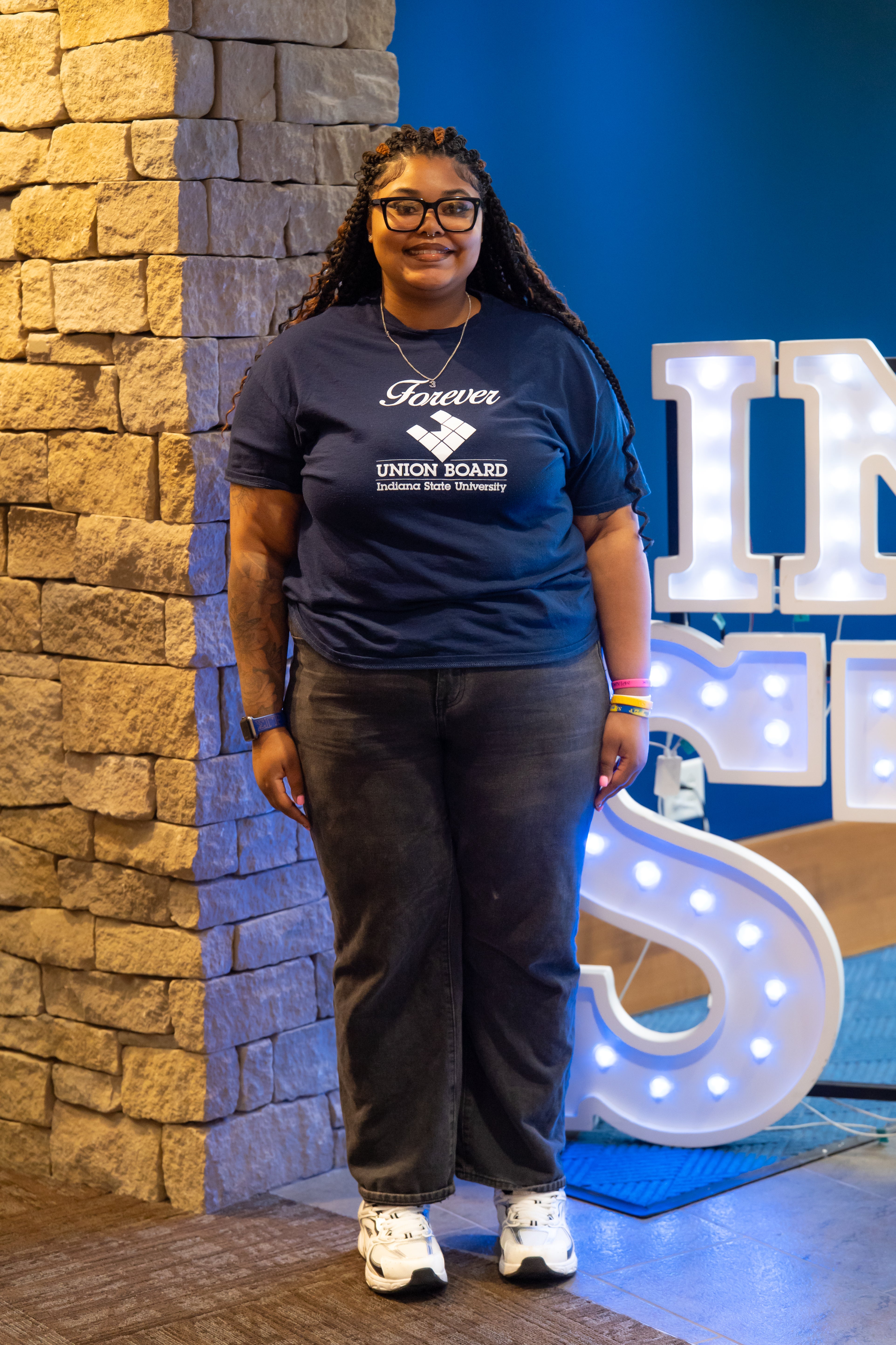 A smiling person with long braided hair and glasses stands indoors beside a stone wall and illuminated “Indiana State” display letters. They are wearing a navy blue Indiana State Union Board T-shirt, dark jeans, and white sneakers, with bracelets and a smartwatch visible on their wrists. Blue walls and lit signage are visible in the background.