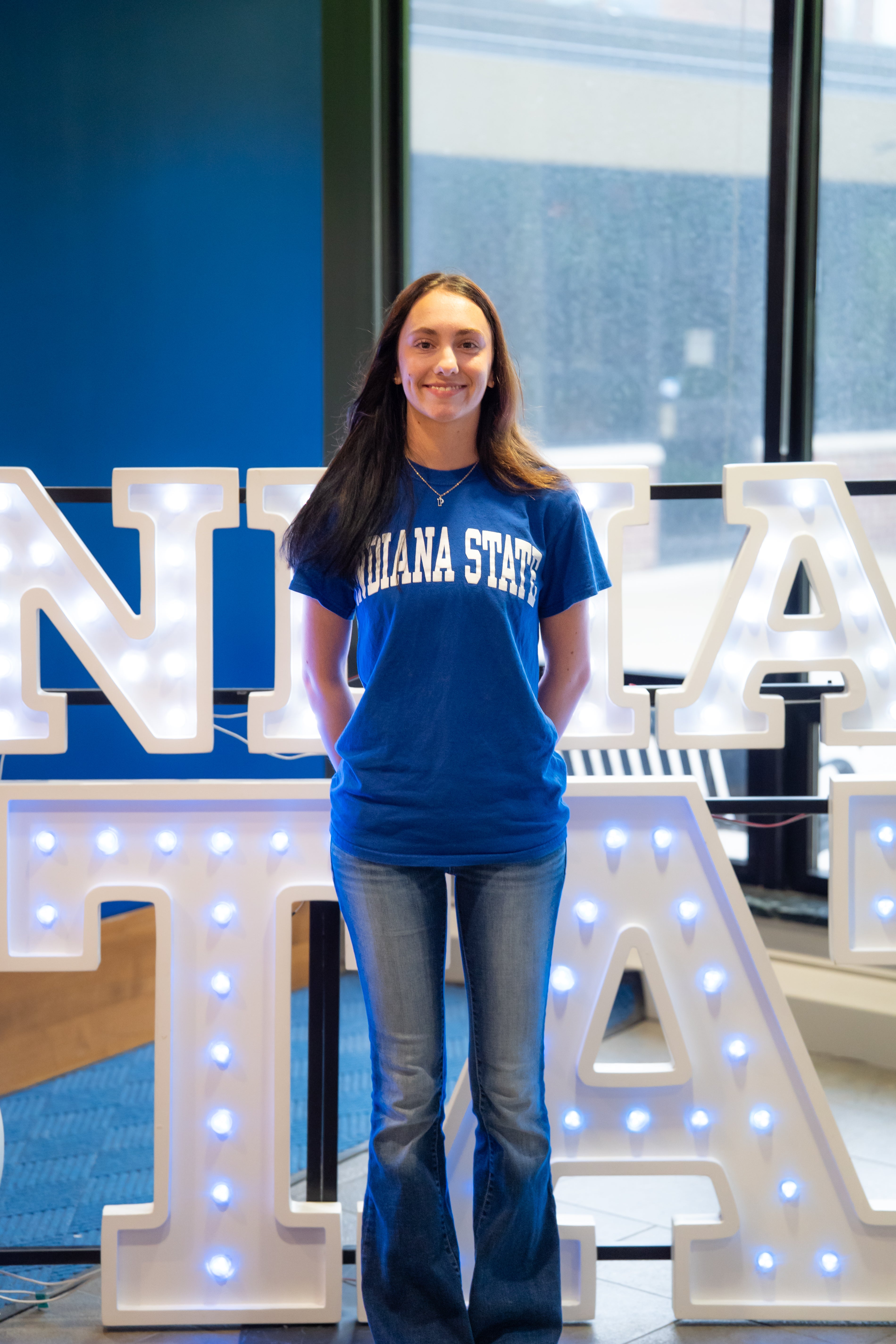 A smiling person with long dark hair stands indoors in front of illuminated “Indiana State” display letters and large windows. They are wearing a bright blue Indiana State T-shirt, blue jeans, and a necklace, with their hands behind their back. Blue walls and lit signage are visible in the background.