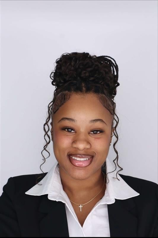 A smiling person with curly hair styled in an updo poses against a light gray background. They are wearing a black blazer over a white collared shirt, along with a necklace featuring a cross pendant.