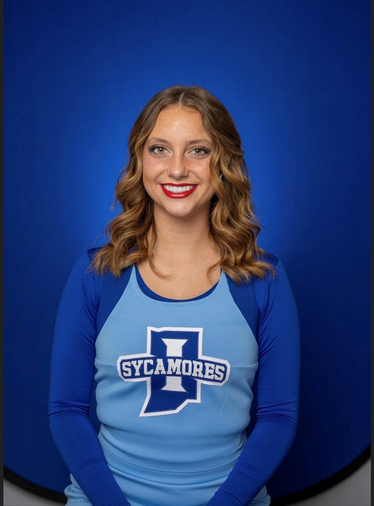 A smiling person with shoulder-length wavy light brown hair poses against a solid blue background. They are wearing a light blue and royal blue Sycamores top with long sleeves and bright red lipstick.