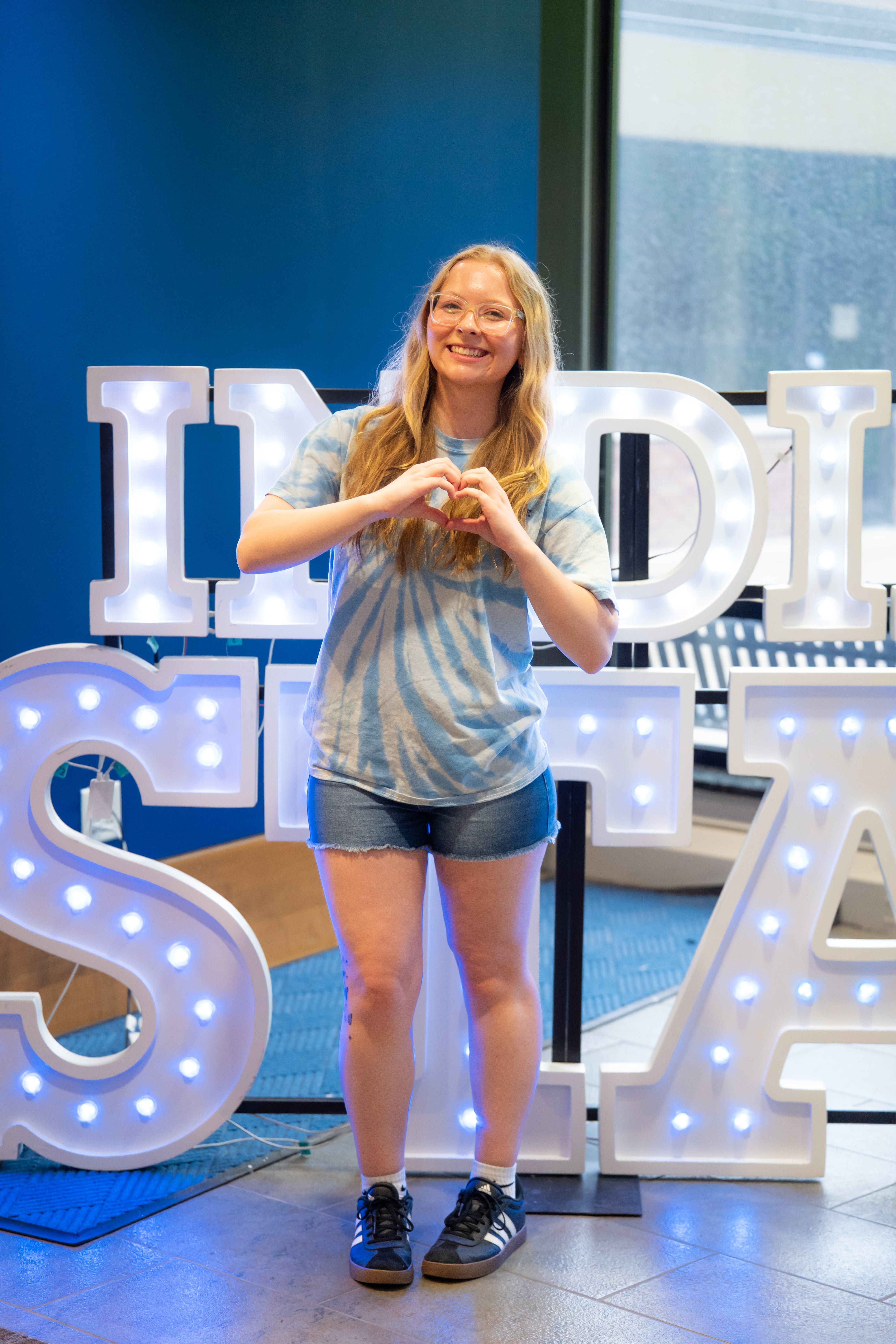 A smiling person with long blonde hair and glasses stands indoors in front of illuminated “Indiana State” display letters and large windows, making a heart shape with their hands. They are wearing a light blue tie-dye T-shirt, denim shorts, white socks, and black sneakers. Blue walls and lit signage are visible in the background.