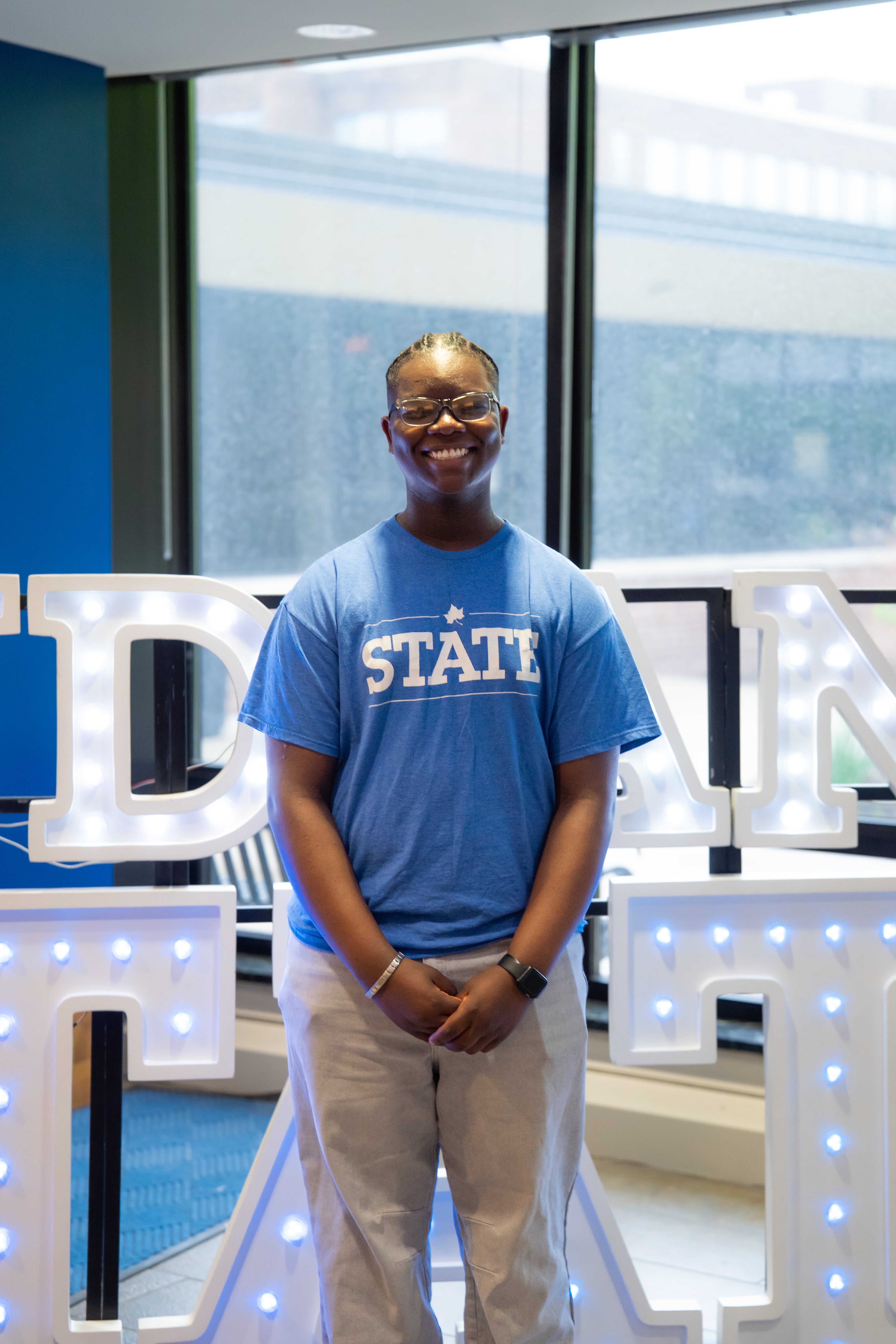 A smiling person with short braided hair and glasses stands indoors in front of illuminated “Indiana State” display letters and large windows. They are wearing a bright blue “STATE” T-shirt, light-colored pants, and a smartwatch, with their hands clasped in front of them. Blue walls and lit signage are visible in the background.
