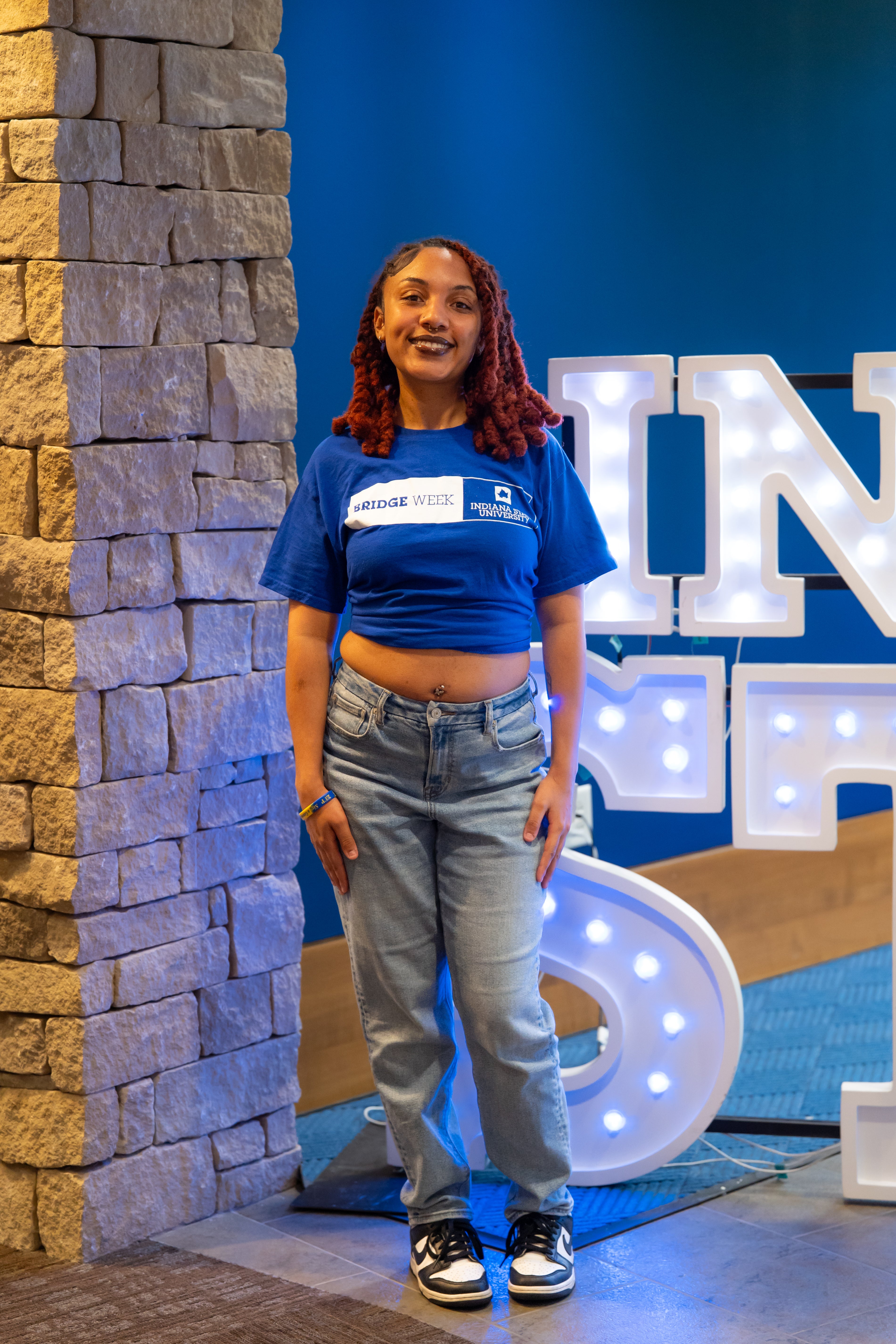 A smiling person with shoulder-length red braided hair stands indoors beside a stone wall and illuminated “Indiana State” display letters. They are wearing a blue Indiana University Bridge Week cropped T-shirt, light blue jeans, and black-and-white sneakers, with a visible belly button piercing and dark lipstick. Blue walls and lit signage are visible in the background.