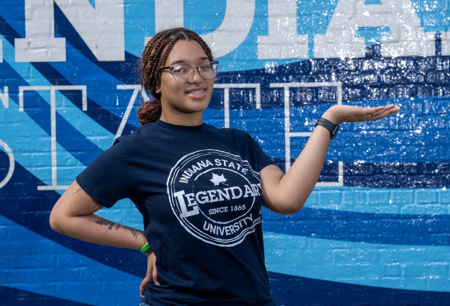 A person with braided hair, glasses, and a septum piercing poses in front of a blue Indiana State mural wall. They are wearing a dark Indiana State University “Legendary Since 1865” T-shirt and a smartwatch, with one hand on their hip and the other raised outward in a presenting gesture. Tattoos are visible on one forearm.