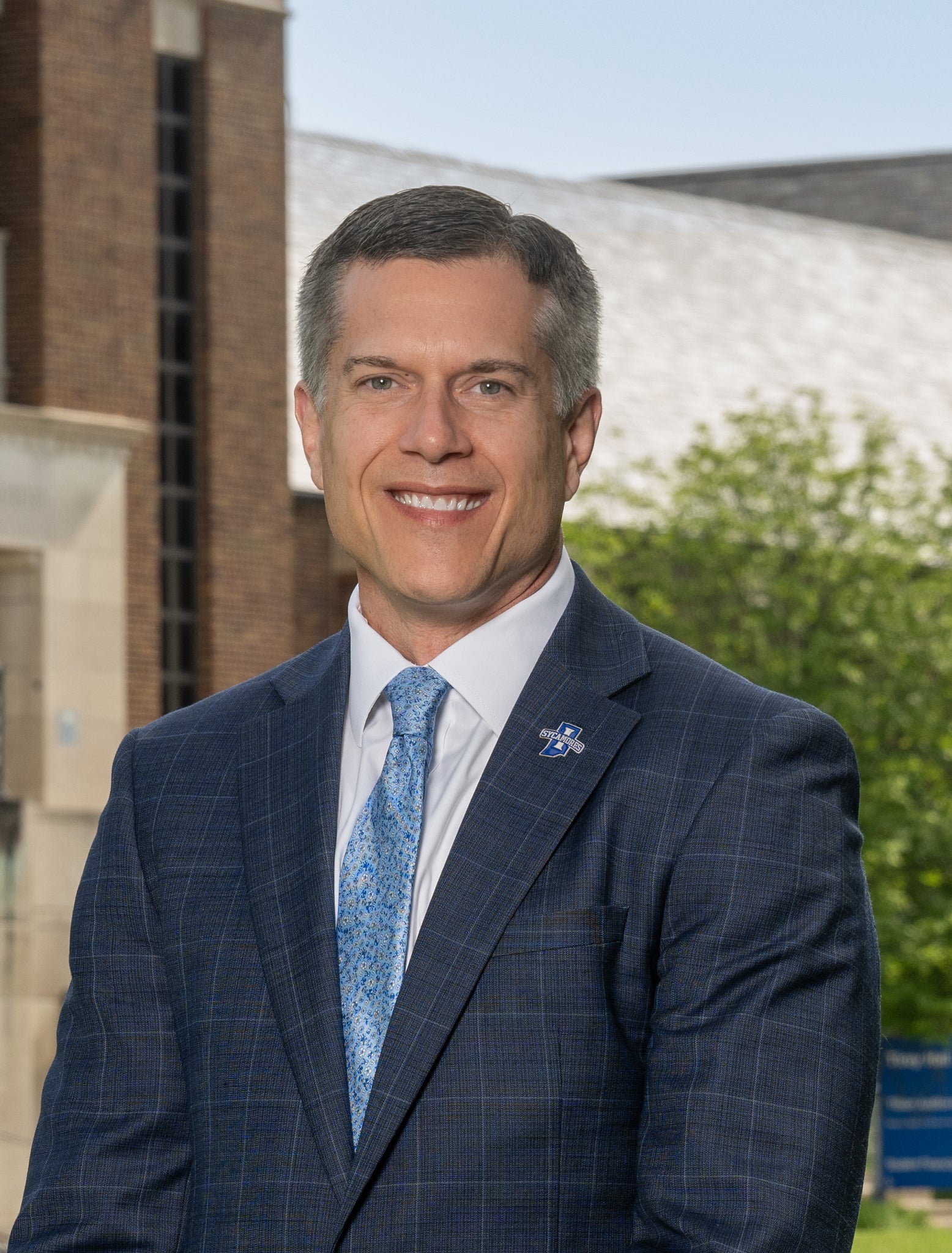 The image shows a professionally dressed man in a dark blue suit with a light blue patterned tie, standing outdoors in front of campus-style buildings and greenery. He is wearing a lapel pin with the Indiana State University Sycamores logo. The photo appears to be an official university headshot or leadership portrait.