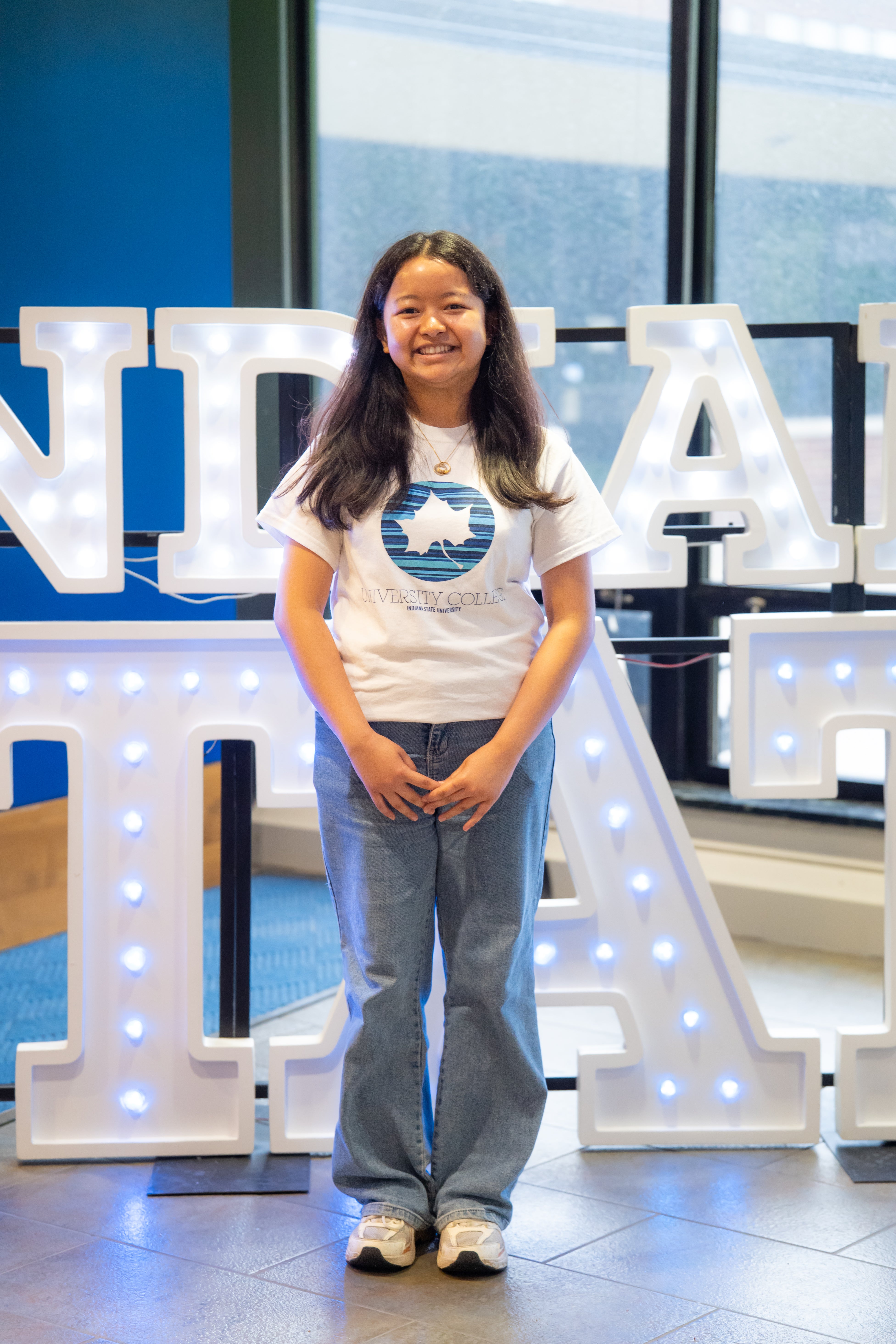 A smiling person with long dark hair stands indoors in front of illuminated “Indiana State” display letters and large windows. They are wearing a white Indiana State University College T-shirt, light blue jeans, white sneakers, and a necklace, with their hands clasped in front of them. Blue walls and lit signage are visible in the background.