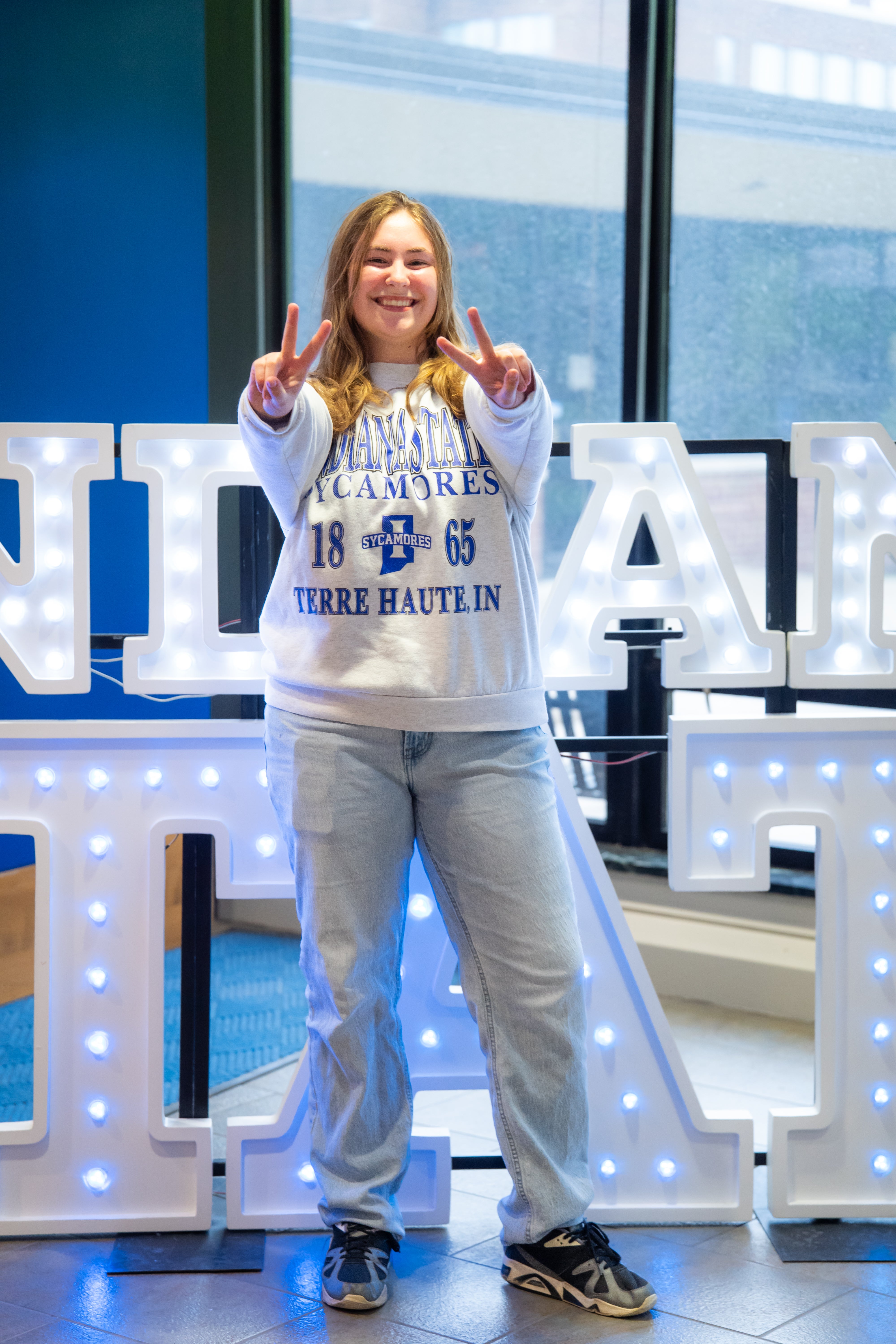 A smiling person with shoulder-length light brown hair stands indoors in front of illuminated “Indiana State” display letters and large windows, holding up peace signs with both hands. They are wearing a white Indiana State Sycamores sweatshirt, light blue jeans, and black-and-gray sneakers. Blue walls and lit signage are visible in the background.