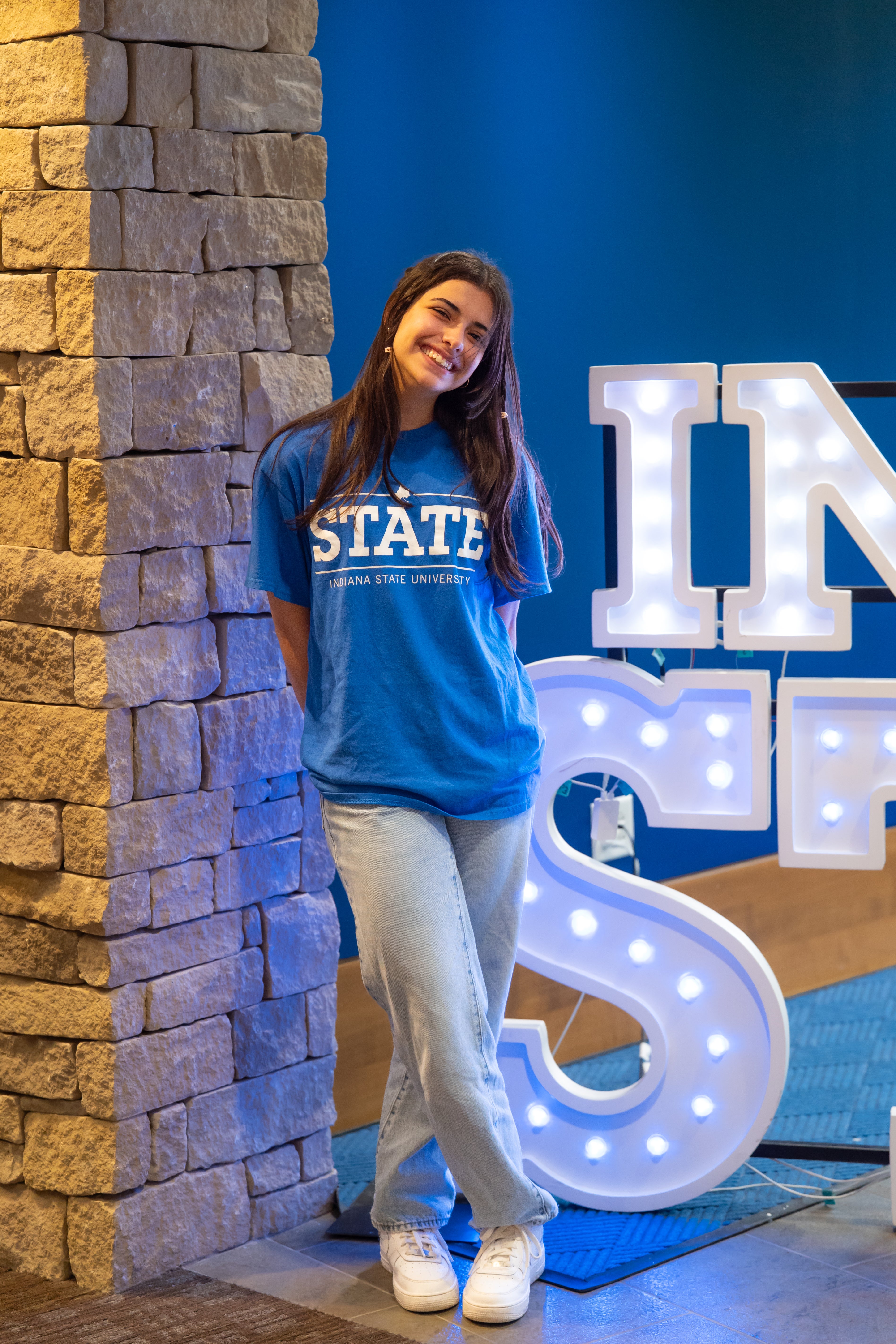 A smiling person with long dark hair stands indoors beside a stone wall and illuminated “Indiana State” display letters. They are wearing a bright blue Indiana State University “STATE” T-shirt, light blue jeans, and white sneakers, with their hands behind their back and one leg crossed in front of the other. Blue walls and lit signage are visible in the background.