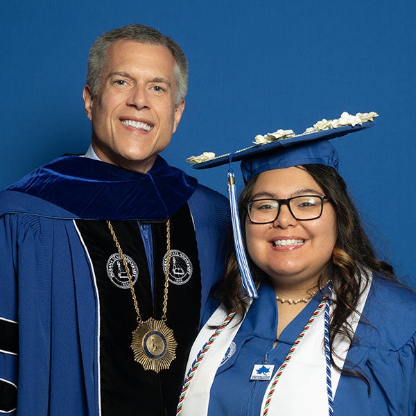 Graduate in cap and gown stands beside Indiana State University President Mike Godard, who is dressed in academic regalia. Both face the camera against a blue studio background. The graduate wears glasses, a decorated mortarboard, honor cords, and a stole, while President Godard wears a ceremonial medallion and gown.