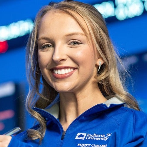 Close-up portrait of a person wearing a blue Indiana State University quarter-zip jacket with “Scott College” text visible on the chest, smiling toward the camera against a softly blurred blue background with lights.