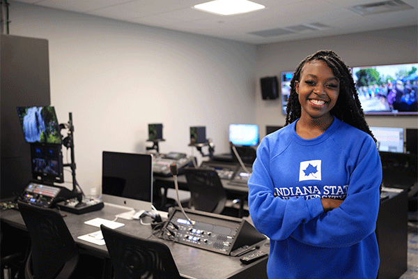 Student standing in a multimedia production studio with video editing and broadcast equipment.