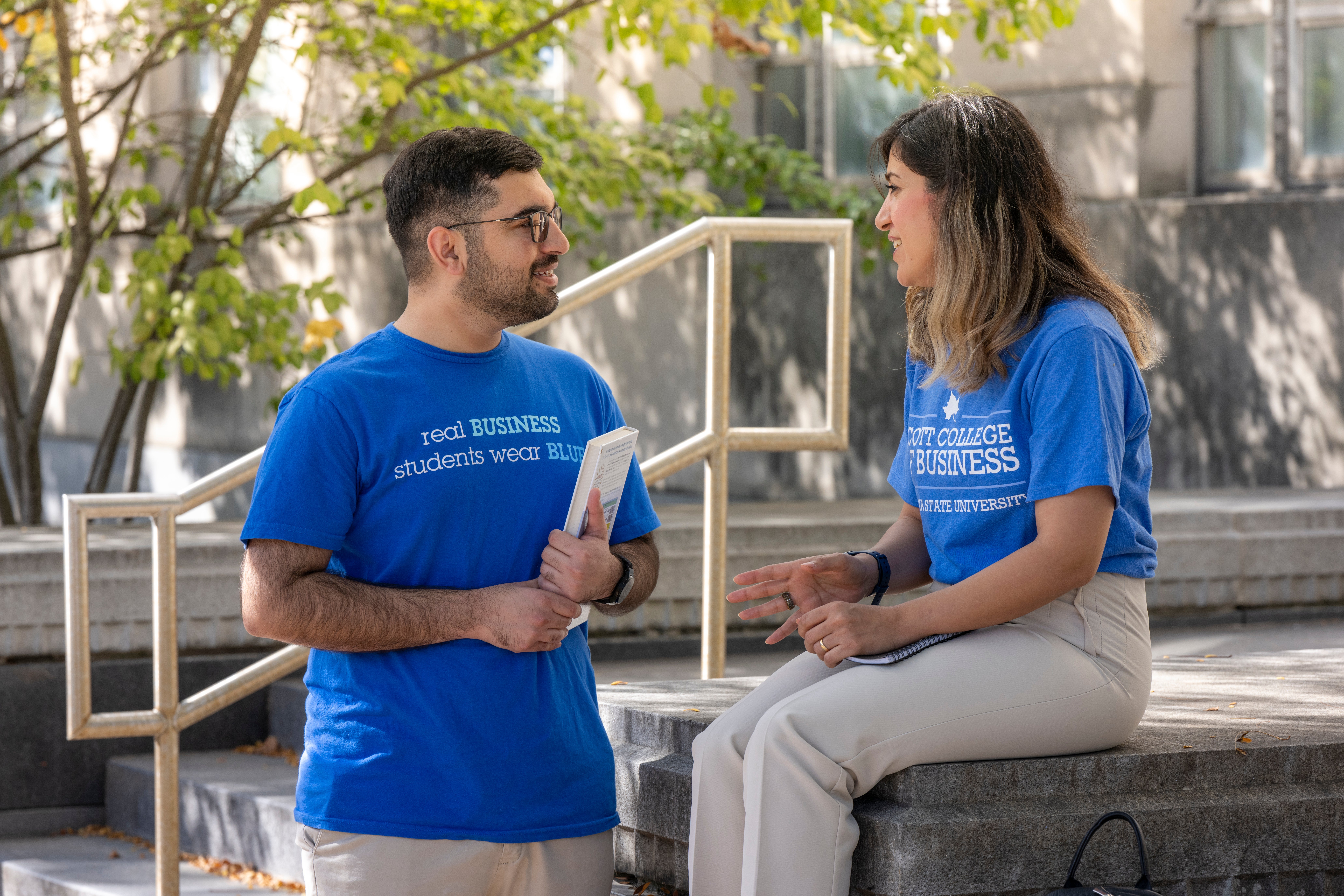 Two people in blue shirts converse outdoors on a sunny day. One person's shirt reads, "Real Business Students Wear Blue." The other person's shirt says, "Scott College of Business." They are near steps and surrounded by trees. One holds a notebook, and the other is seated on a stone surface.