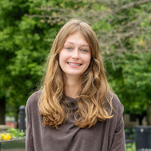 Person with long hair wearing a brown shirt, standing outdoors on a campus walkway with trees and flowers in the background.