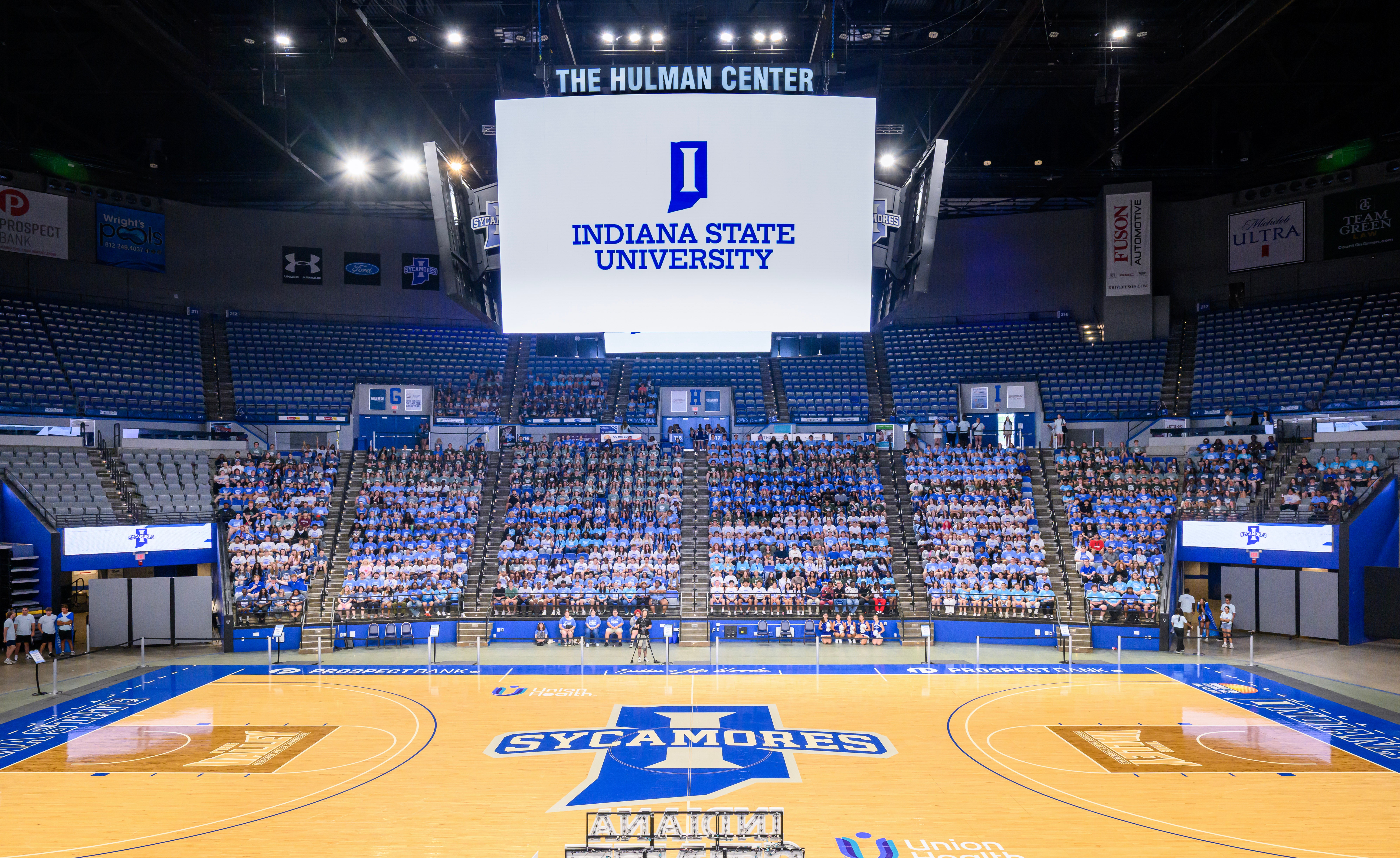 The image shows the interior of the Hulman Center at Indiana State University. It features a large basketball court with the Sycamores logo at center court. A big screen above displays the university's logo and name. The seating area is filled with people, most wearing white shirts.