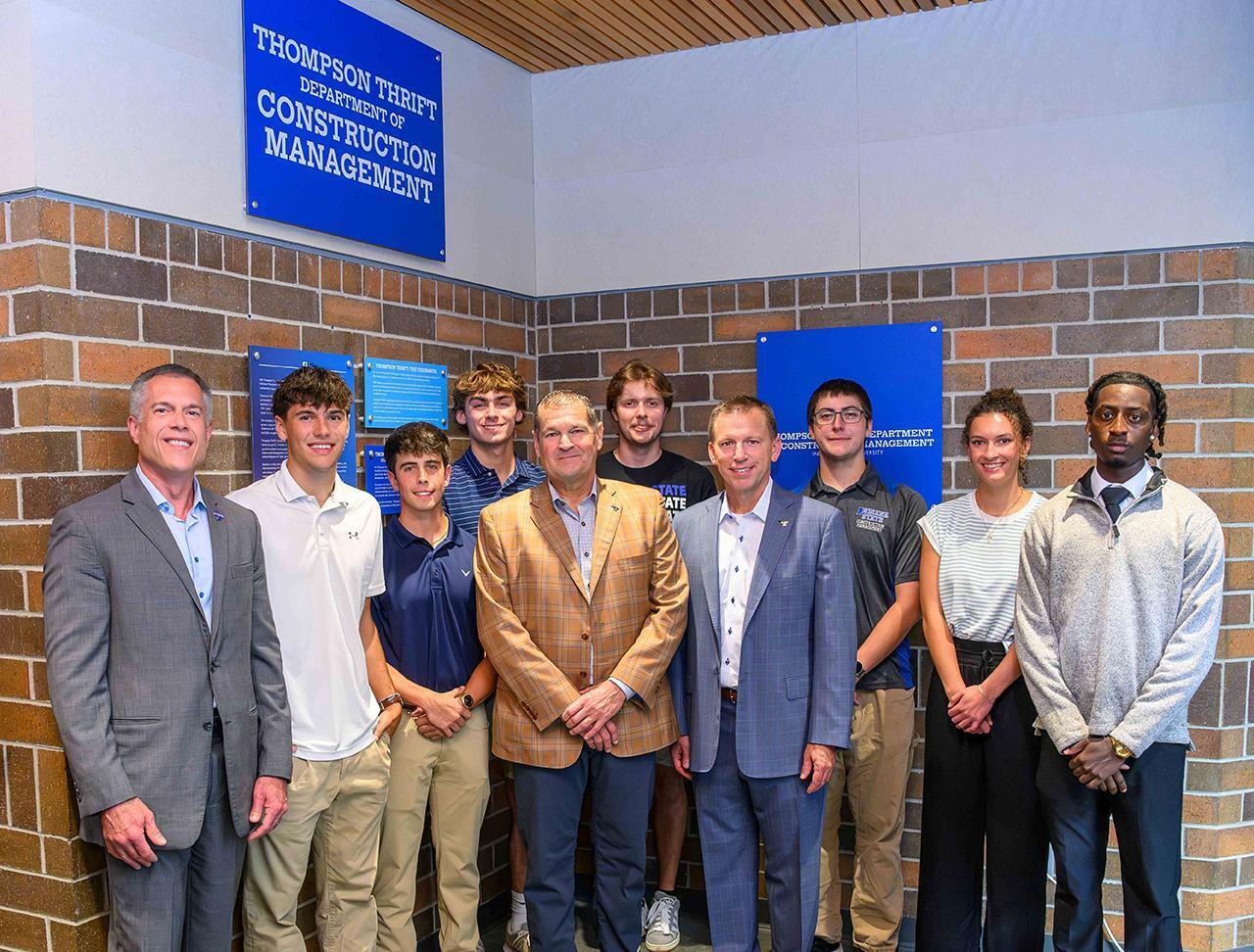 A group of nine people stand in front of a brick wall with two blue signs. The signs read "Thompson Thrift Department of Construction Management." They are dressed in formal and semi-formal attire, posing for a group photo.