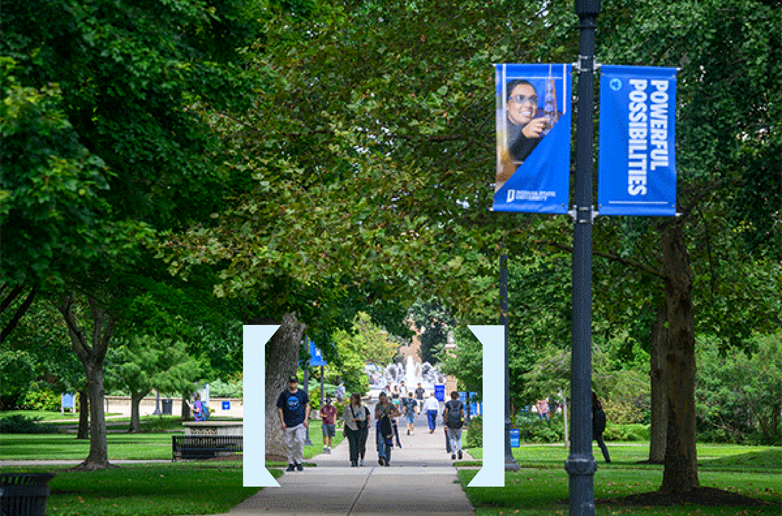 Tree-lined campus walkway with several people walking at a distance along a paved path. Large bracket-shaped graphic elements highlight the center of the walkway, and a blue campus banner on a lamppost reads “POWERFUL POSSIBILITIES.” A fountain is visible in the distance.
