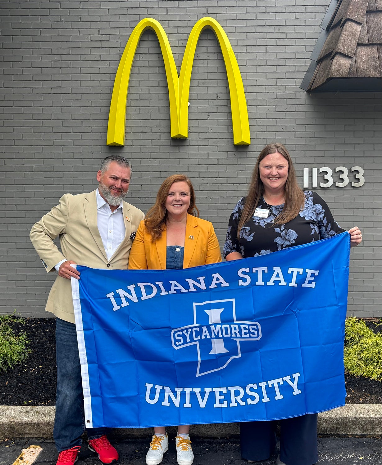 An image features three individuals holding a large, blue "Indiana State University Sycamores" flag in front of a grey brick building adorned with a prominent yellow McDonald's logo.
