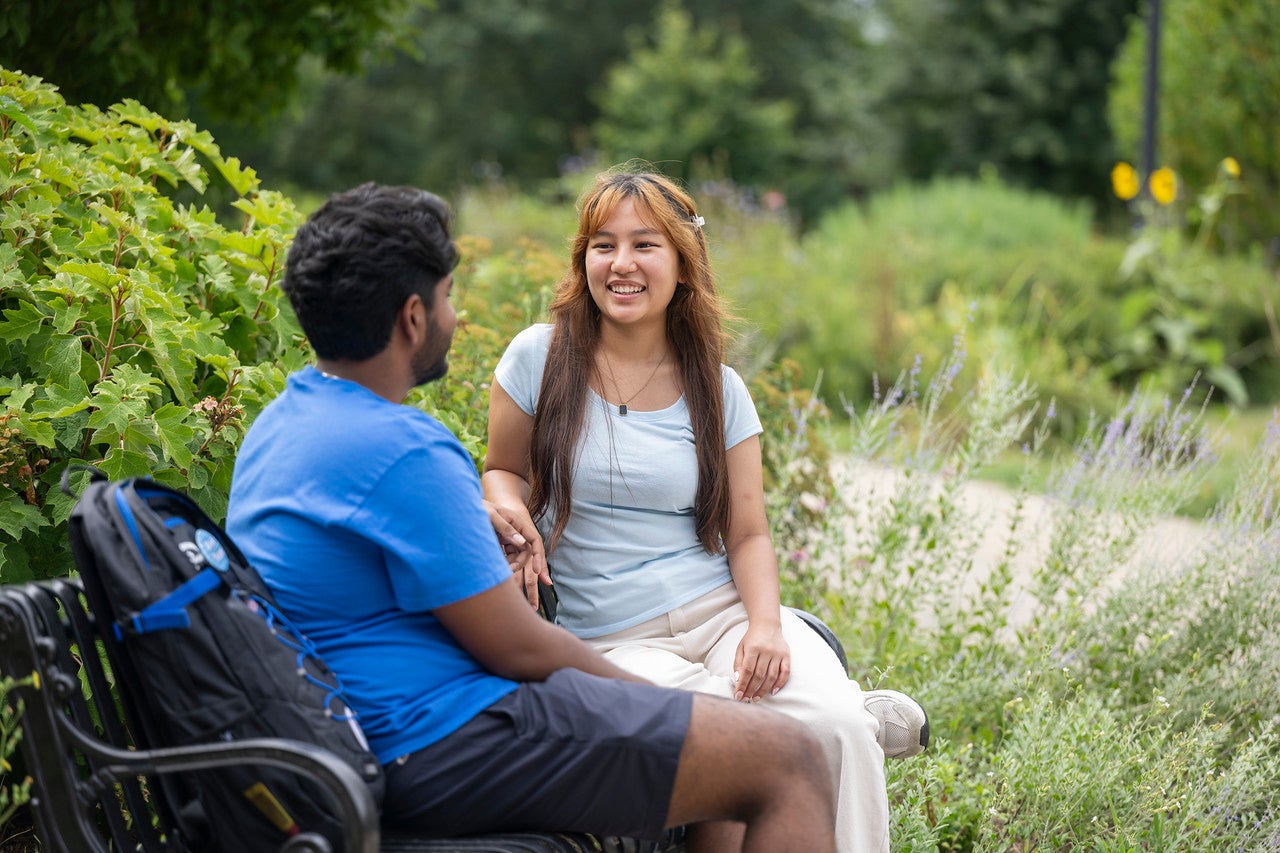 Two students sit on a bench along a landscaped campus path, talking outdoors among trees, shrubs, and flowering plants. A backpack rests on the bench beside one student.