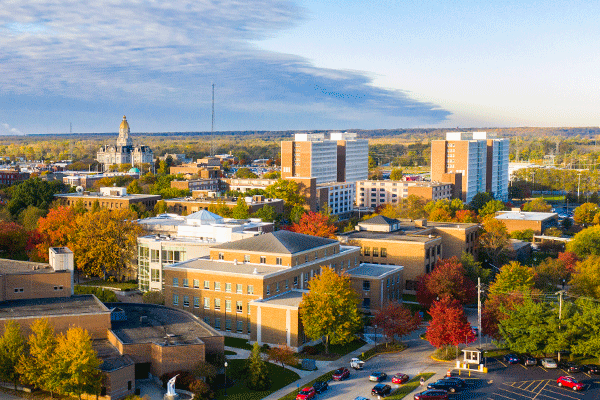 Aerial view of the Indiana State University campus and surrounding city during autumn. Brick academic buildings, residence halls, streets, and parking areas are surrounded by trees with red, orange, yellow, and green foliage under a partly cloudy sky.