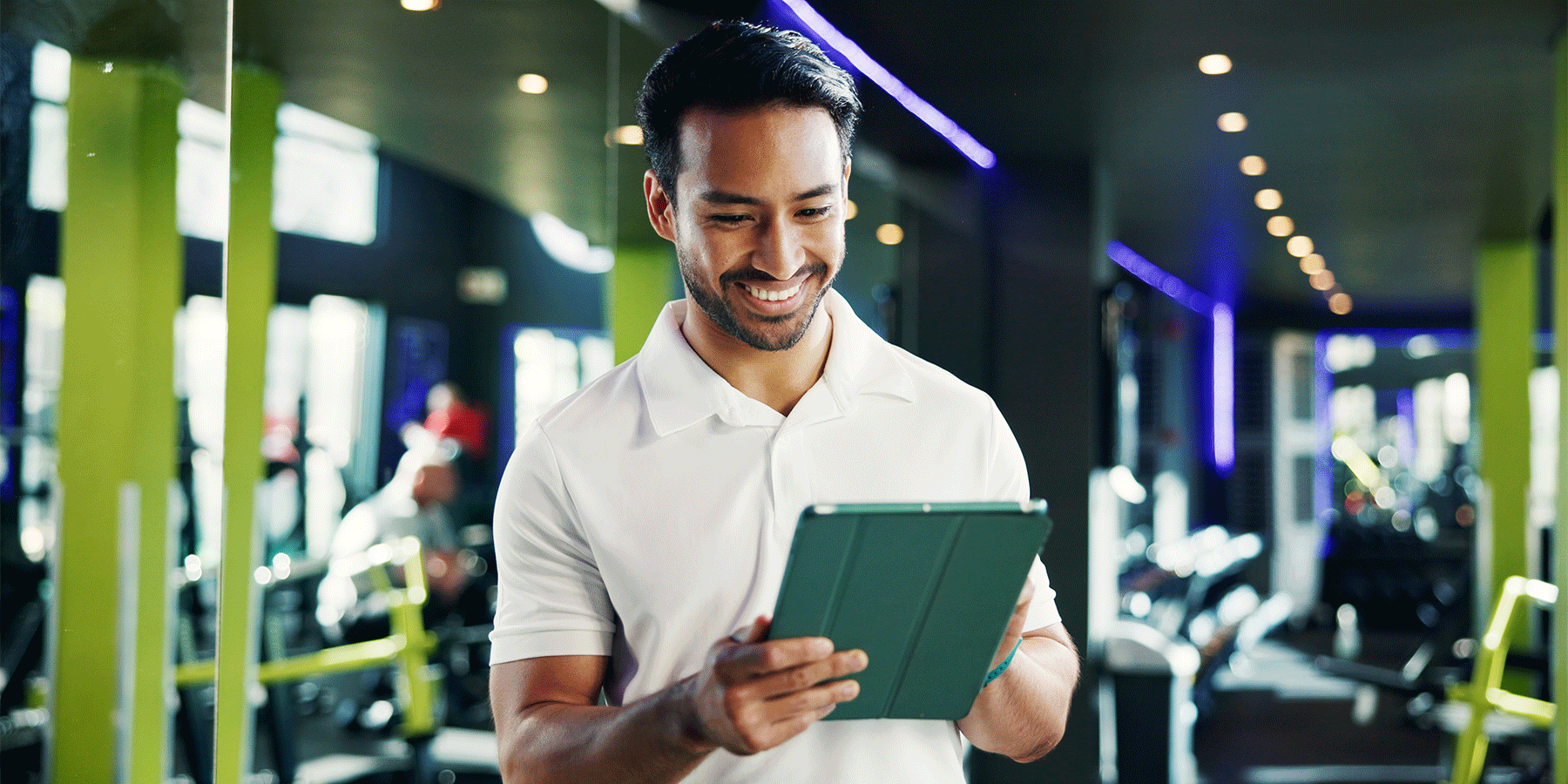 Person standing in a fitness center holding a tablet and looking at the screen. Exercise equipment, mirrors, and bright green columns are visible in the background, with overhead lights reflecting throughout the gym.