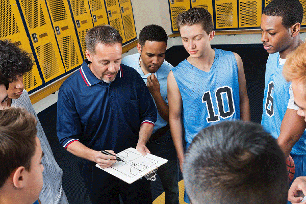 Coach holding a clipboard with a basketball court diagram while standing in a locker room surrounded by players in practice jerseys. Yellow lockers line the walls as the group gathers closely to review the coach’s instructions.