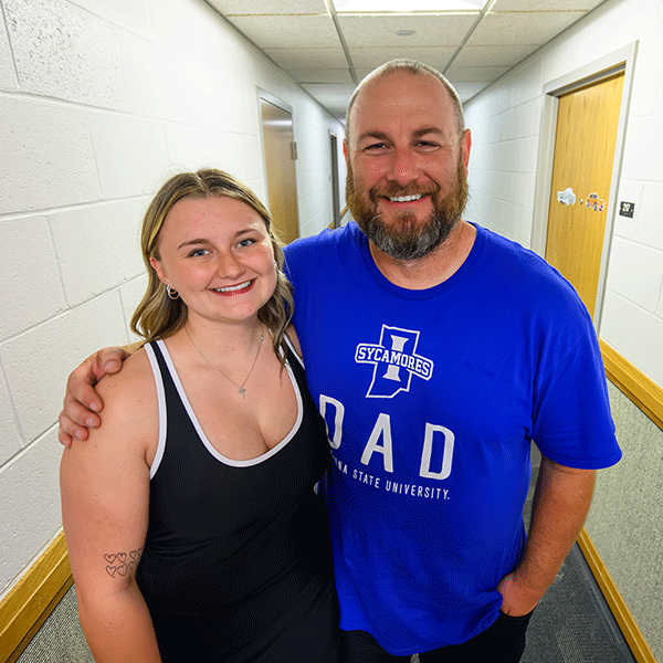 A father and daughter stand together in a residence building hallway, facing the camera. The daughter wears a black tank top, and the dad wears a blue Indiana State University "Sycamores Dad" T-shirt. The hallway has white walls, wood trim, and doors on both sides.