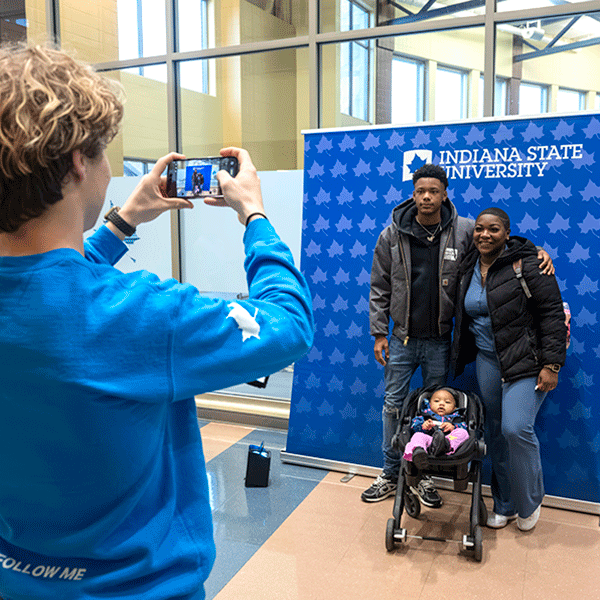 A person in a blue sweatshirt takes a photo of a family standing in front of an Indiana State University backdrop inside a campus building. The group includes two adults and a child in a stroller, posing for the picture in a bright lobby with large windows.