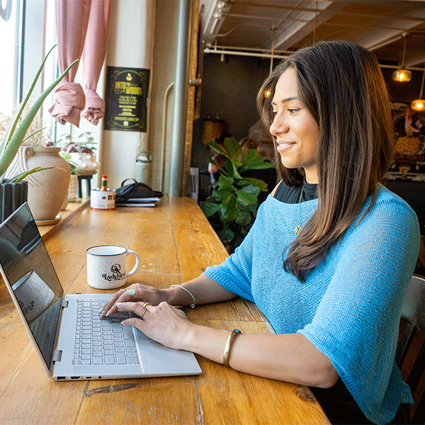 A smiling woman with long, dark hair and wearing a blue top works on a laptop while seated at a long wooden counter inside a café. Large potted plants line the window to the left, allowing natural light to illuminate the workspace. A white mug sits next to the laptop, and additional plants, seating, and hanging lights are visible in the background.