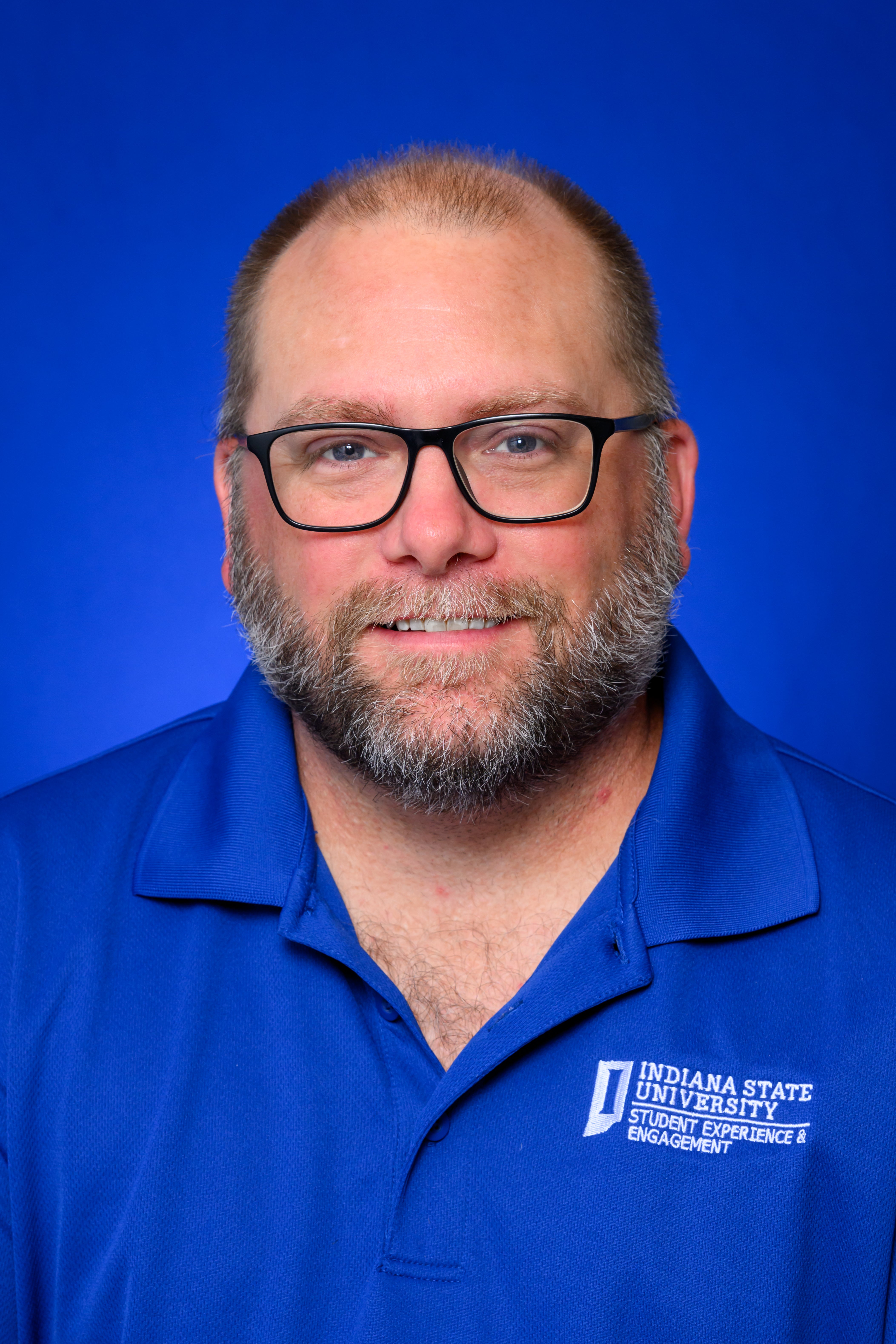 A person with glasses and a beard is smiling against a blue background. They are wearing a blue polo shirt with the Indiana State University Student Experience and Engagement logo.