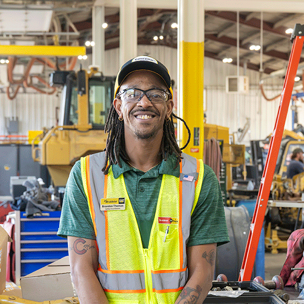 A person wearing safety glasses, a high-visibility safety vest, and a Caterpillar-branded cap stands in a heavy equipment maintenance facility. Construction equipment, a ladder, tools, and workstations are visible throughout the shop in the background.