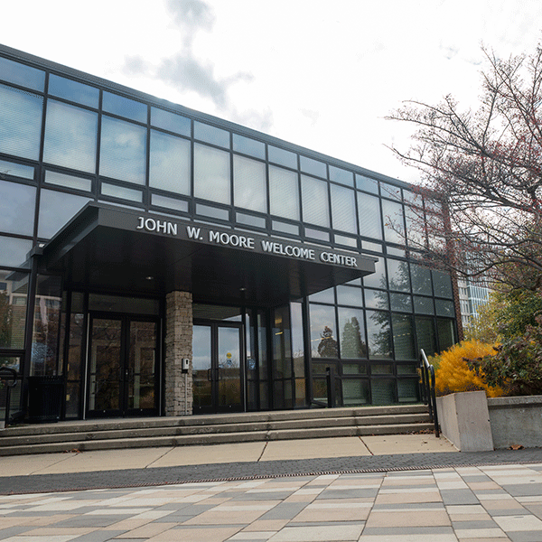 Front entrance of the John W. Moore Welcome Center, a modern glass-front building with a covered entryway, steps leading to glass doors, and trees and landscaping on both sides of the walkway.